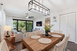 A modern living room with a wooden dining table, woven placemats, a ceiling fan, and a view of the outdoors. - D-225 - End Unit - Serene Views -Updated! (Naples)