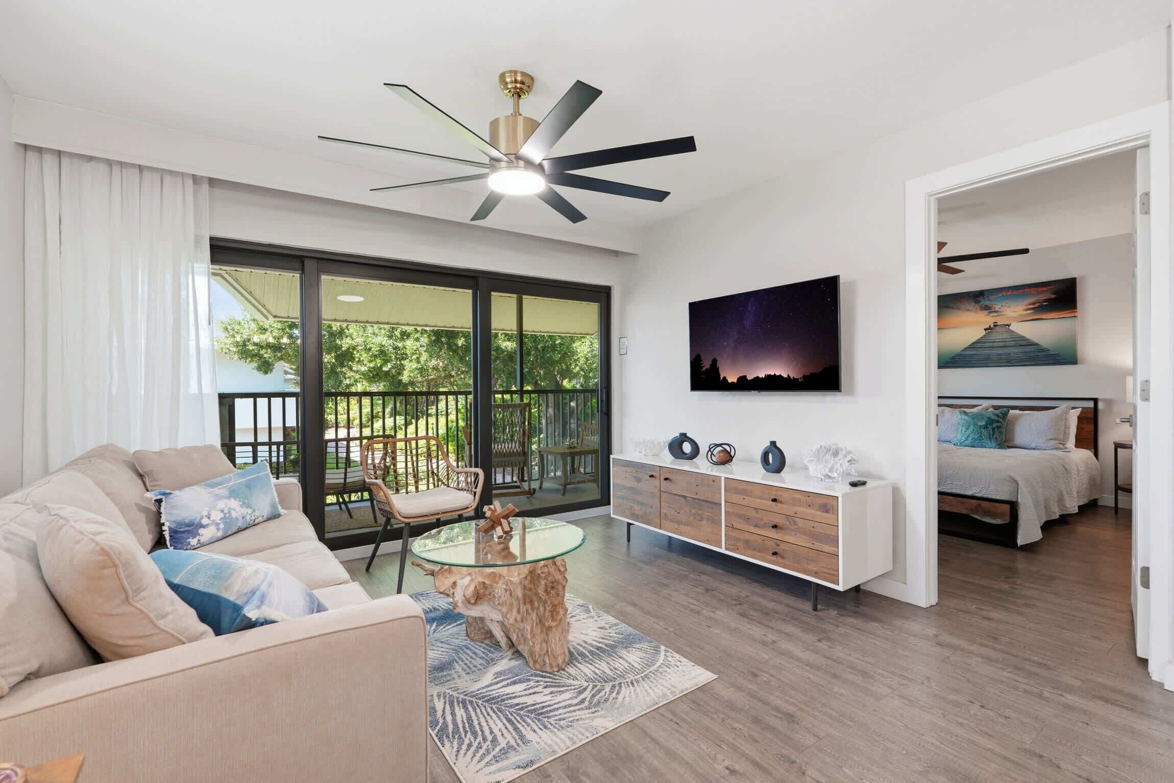 A modern living room with a glass coffee table, a sofa, a dining table, a ceiling fan, and a bed in the adjacent room.