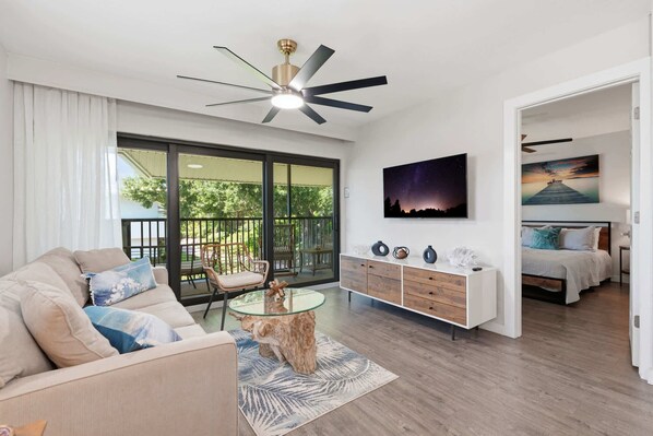 A modern living room with a glass coffee table, a sofa, a dining table, a ceiling fan, and a bed in the adjacent room. - D-225 - End Unit - Serene Views -Updated! (Naples)