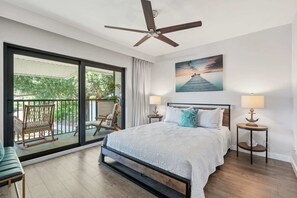 A bedroom with a bed, two bedside tables, a ceiling fan, a large window with a view of greenery, and a painting on the wall. - D-225 - End Unit - Serene Views -Updated! (Naples)