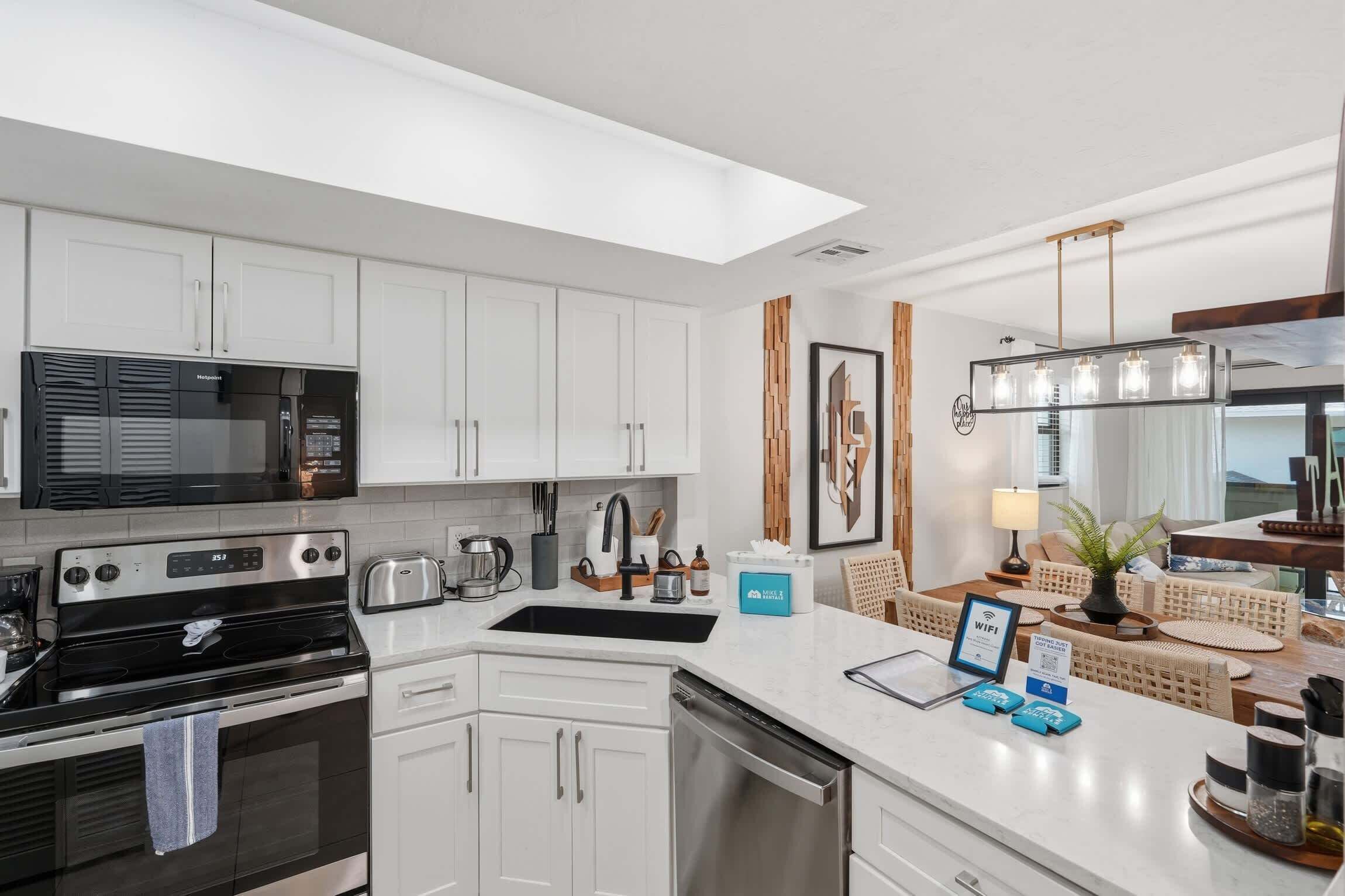 Une cuisine moderne avec des meubles blancs, un four à micro-ondes, une cuisinière et un évier. On y trouve également un coin repas avec une table et des chaises, une plante et un panneau indiquant la présence d’une connexion Wi-Fi.