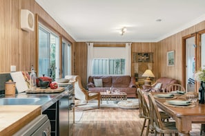 A kitchen with wooden cabinets, a dining area with a table set for two, and a living room with a sofa and a coffee table. - Sugar Shack Currarong (Currarong)