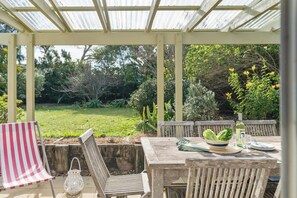 A covered patio with a wooden table set for dining, surrounded by chairs, and a view of a garden. - Sugar Shack Currarong (Currarong)