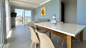 A dining area with a white table, beige chairs, and a vase. There is a painting on the wall and a view through the glass door.