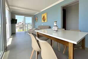 A dining area with a white table, beige chairs, and a vase. There is a painting on the wall and a view through the glass door.