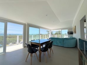 Ein modernes Esszimmer mit Tisch und Stühlen, einem blaugrünen Sofa und großen Fenstern mit Blick auf das Meer und eine ferne Skyline.