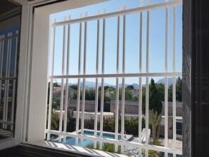 Una ventana con barrotes blancos, una piscina y vistas a casas y montañas.