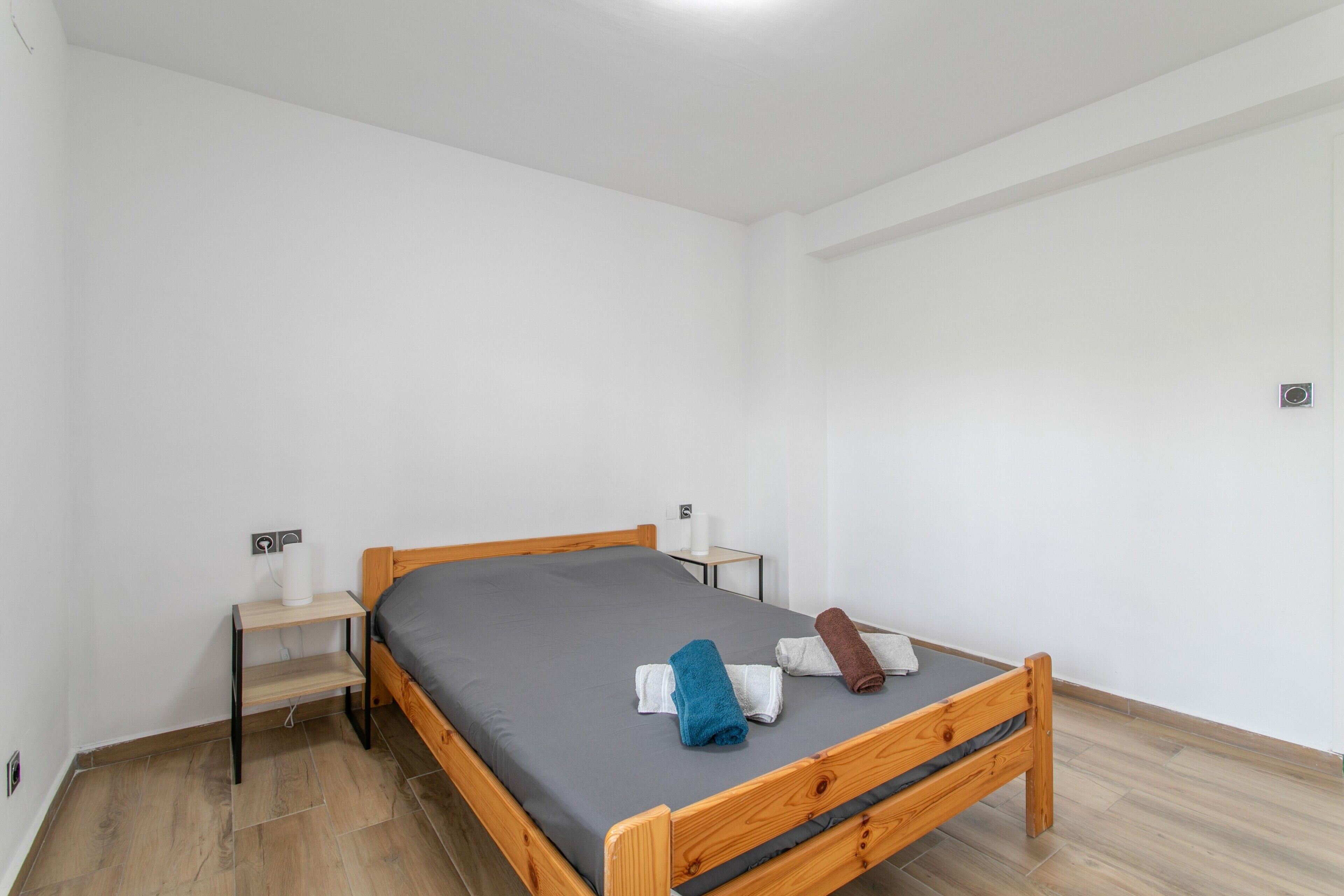A single bed with a wooden frame and grey bedding in a minimalistic room with white walls and a wooden floor.