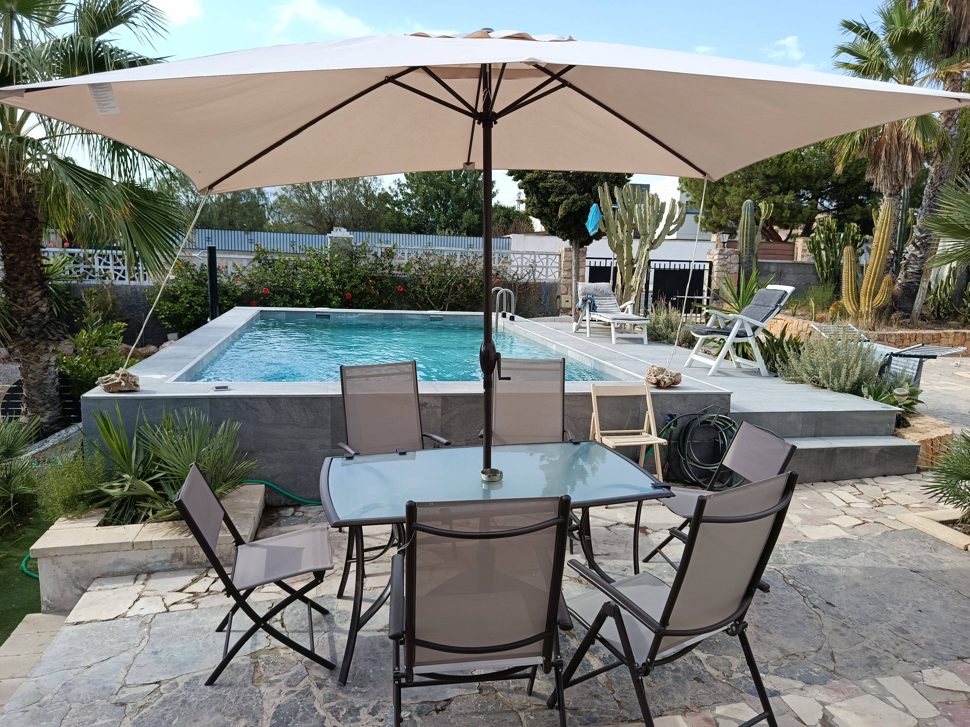 A poolside area with a table and chairs under a large umbrella, surrounded by cacti and palm trees.
