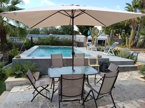 A poolside area with a table and chairs under a large umbrella, surrounded by cacti and palm trees.