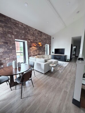 Una sala de estar moderna con una pared de acento de piedra, un conjunto de sofás, un comedor con mesa y sillas de madera, y un televisor.