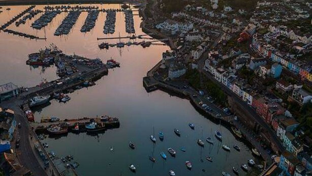Vista aérea de un pueblo costero con un puerto deportivo, edificios y un atardecer.