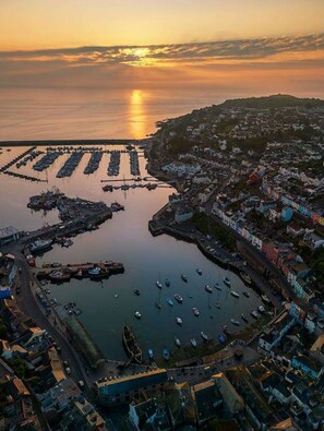 Vista aérea de un pueblo costero con un puerto deportivo, edificios y un atardecer.
