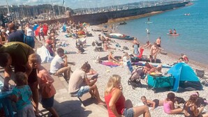 Una playa concurrida con gente tomando el sol y nadando, barcos en el agua y un muelle al fondo.
