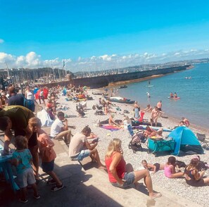 Una playa concurrida con gente tomando el sol y nadando, barcos en el agua y un muelle al fondo.