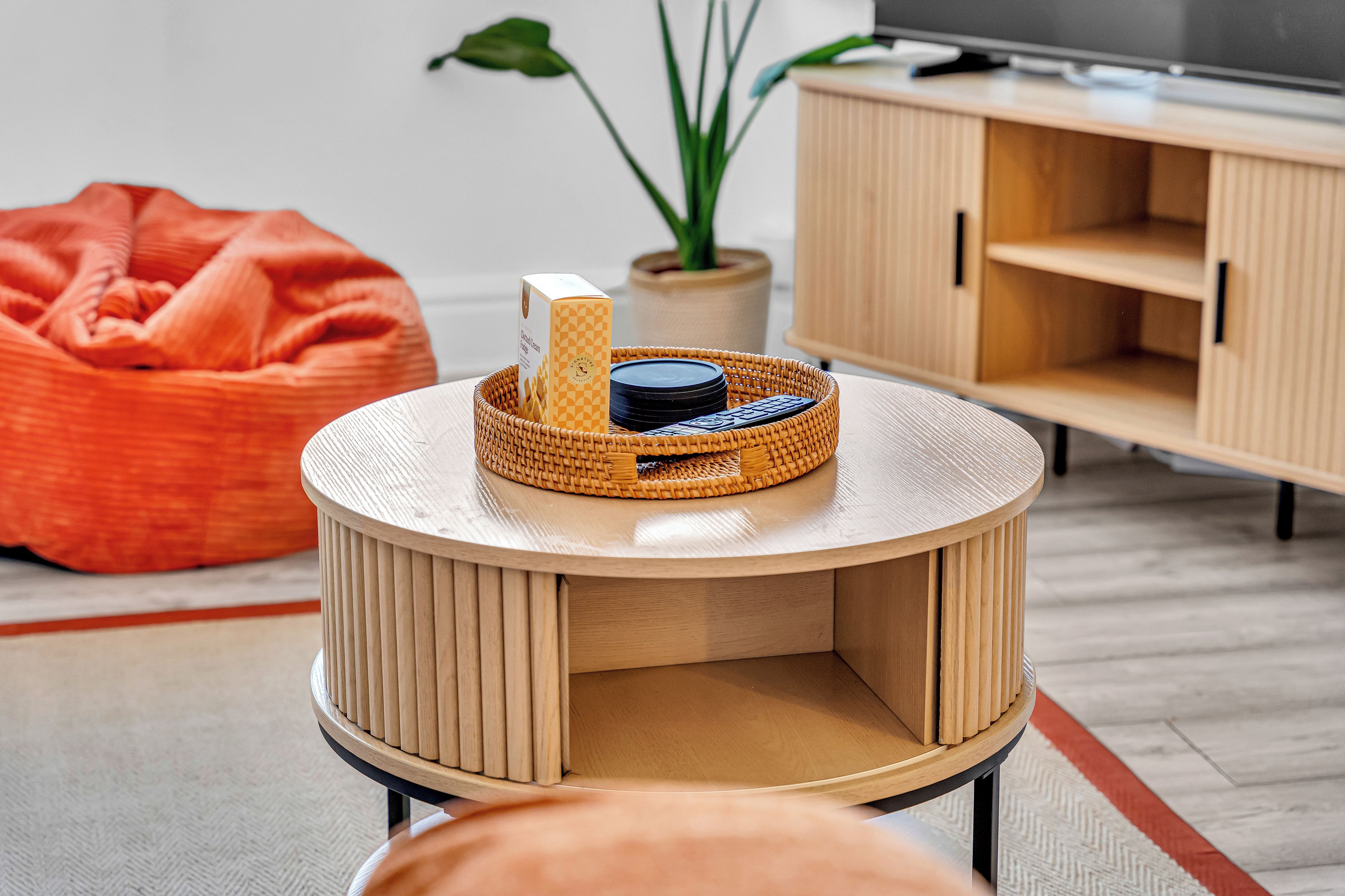 Una mesa de madera redonda con una cesta tejida que contiene un control remoto y una caja, un sillón orejero rojo y un mueble de madera con un televisor al fondo.