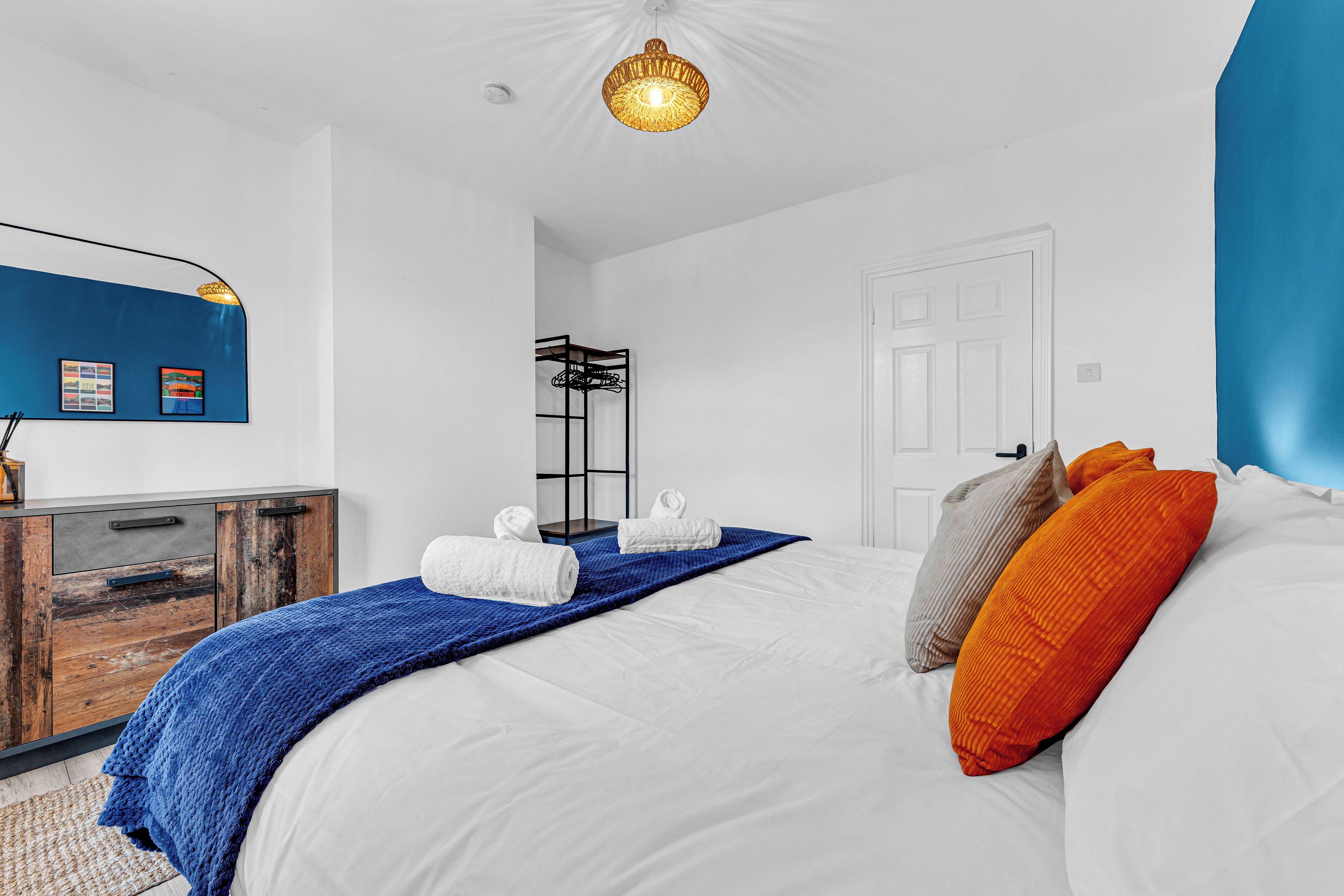 Habitación de hotel con cama, manta azul, toallas blancas, cómoda de madera y lámpara fijada a la pared.