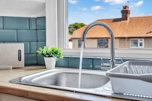 Un lavamanos con agua corriente, una planta en maceta y una ventana con vista a una casa.