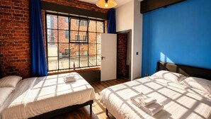 Quarto com duas camas, parede de tijolos, uma janela grande com cortinas azuis e uma parede azul decorativa.