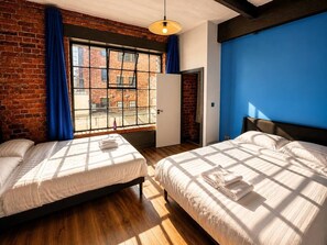 Ein Zimmer mit zwei Betten, einer Ziegelwand, einem großen Fenster mit blauen Vorhängen und einer Wand in Blautönen.