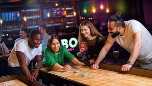 A group of people playing a board game at a bar with a counter and shelves in the background.