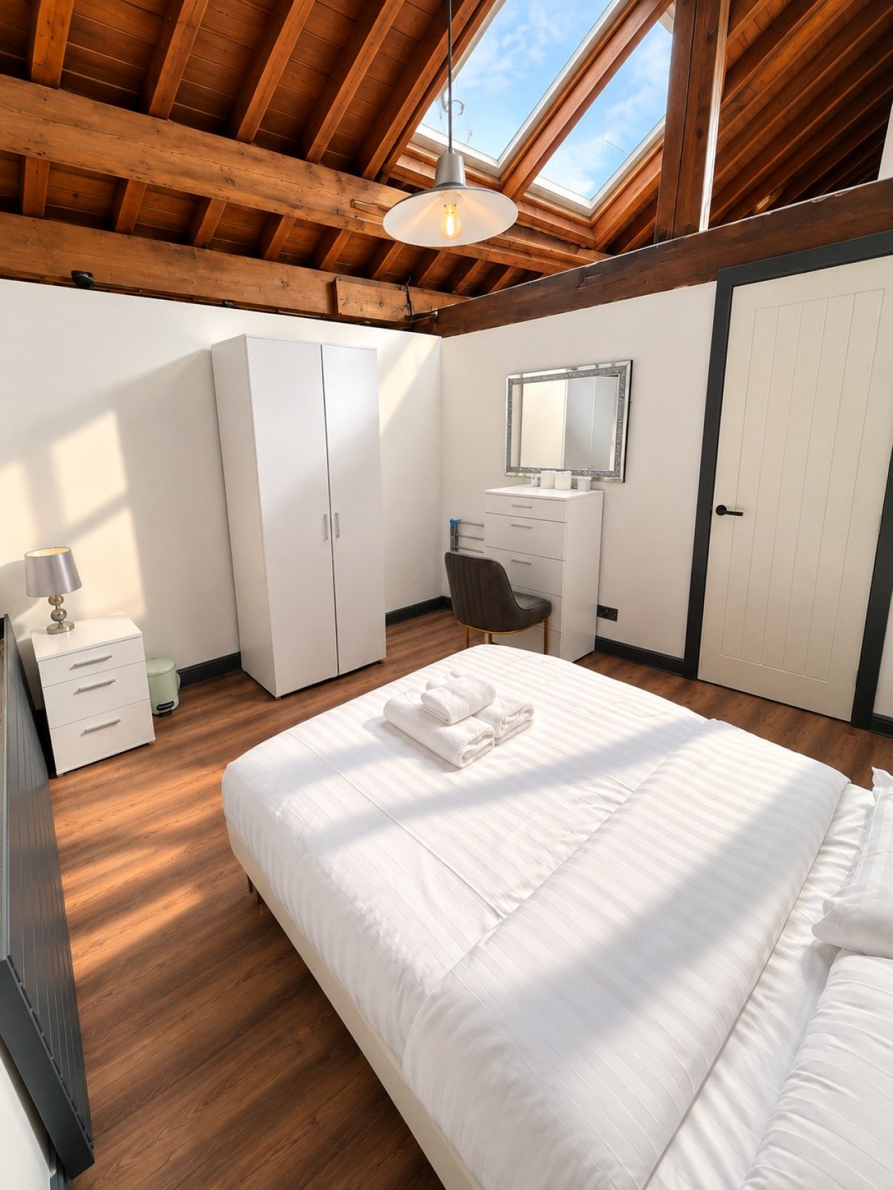 A bedroom with a skylight, a bed with white linens, a desk with a mirror, and a wardrobe.
