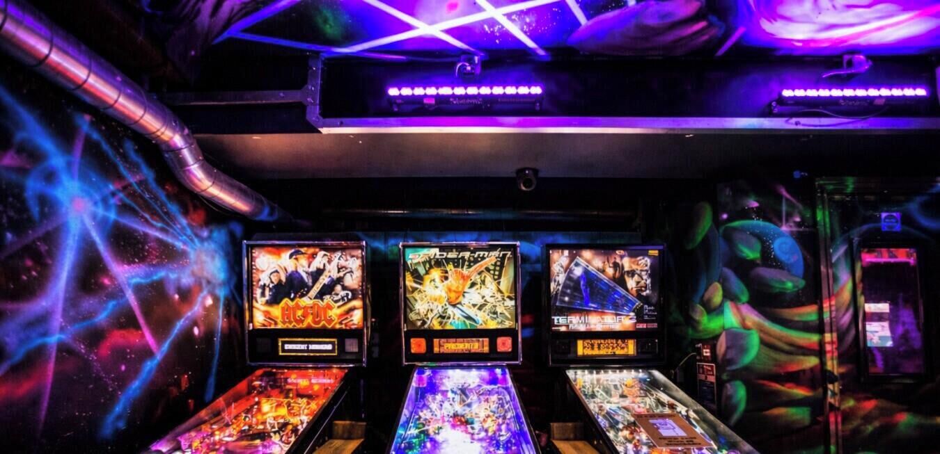 En rad med flipperspel i ett svagt upplyst rum med lila belysning.