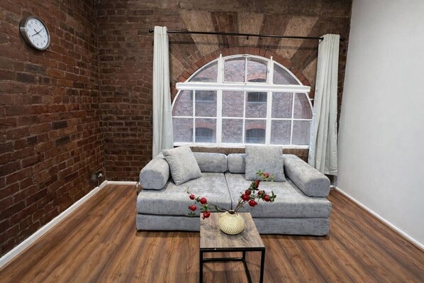 A room with a brick wall, a large window with curtains, a grey sofa, a small table with a vase, and a clock on the wall. - City Centre | Sleeps 6 | Bars, Parking & Netflix (Merseyside)