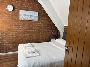 A bedroom with a brick wall, a clock, a framed picture, a bed with white linens, and a wooden door. - City Centre | Sleeps 6 | Bars, Parking & Netflix (Merseyside)