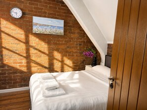 A bedroom with a brick wall, a clock, a painting, a bed with white linens, and a wooden door. - City Centre | Bars & Shops | Free Parking (Merseyside)
