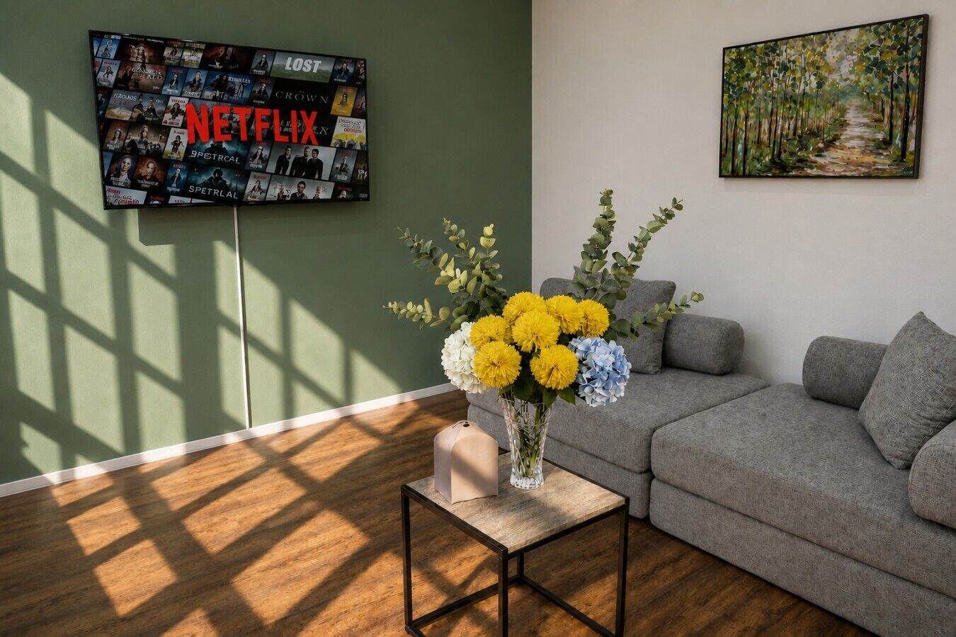 A living room with a grey sofa, a small table with a vase of flowers, and a wall-mounted TV displaying various Netflix shows.