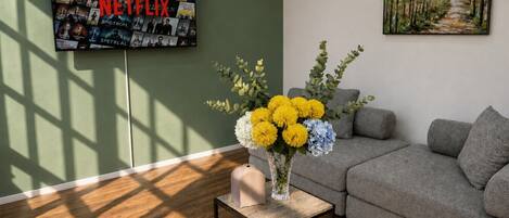 Una sala de estar con un sofá gris, una mesita con un jarrón de flores y un televisor montado en la pared mostrando varias series de Netflix.