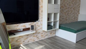 Ein Zimmer mit einer Wand im Steinoptik, einem an der Wand befestigten Fernseher, einem weißen Regal und einem Bett mit einem grünen Topper, sowie einem leuchtend grünen Stuhl.
