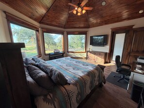 Quarto com cama, escrivaninha, cadeira, televisão e janelas amplas.
