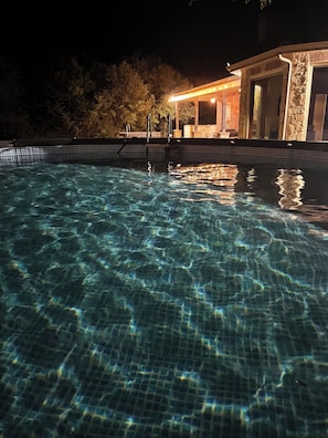 Une piscine aux eaux cristallines, bordée de carreaux, avec une terrasse en bois et, au fond, un bâtiment dont la façade est en pierre.