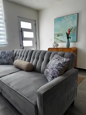 Ein graues Sofa mit gemusterten Kissen, ein Holznachttisch mit einer Vase und ein Bild an der Wand.