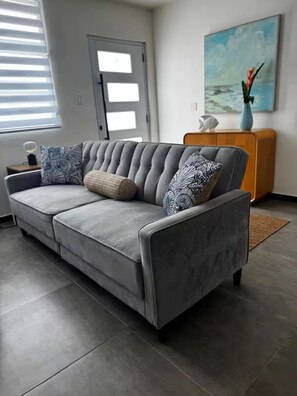 Ein graues Sofa mit gemusterten Kissen, ein kleiner Beistelltisch mit einer Lampe, eine Tür und ein Gemälde an der Wand.