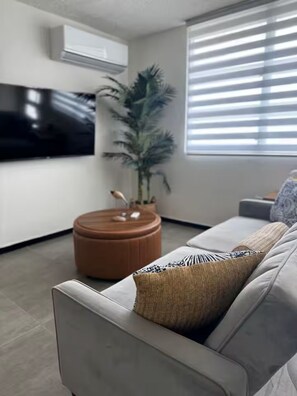 Ein modernes Wohnzimmer mit einem grauen Sofa, einem runden Hocker, einer großen Topfpflanze, einem flachbildfernseher an der Wand und einem Fenster mit Jalousien.