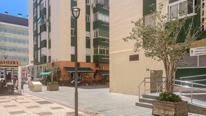 Ein Gebäude mit einer Fassade in Grün- und Beigetönen, einer Außenbestuhlung und einem Baumkübel.