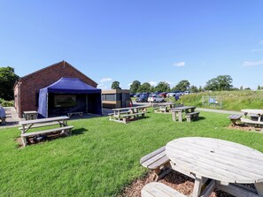 A picnic area with tables and a tent, a brick building, and a parking lot with cars. - Escape II (Northwich)