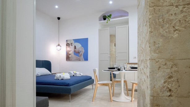 A modern bedroom with a bed, a desk, and a chair. There is a painting on the wall and a plant on the ceiling. - ELORO - Guest Houses (Noto)