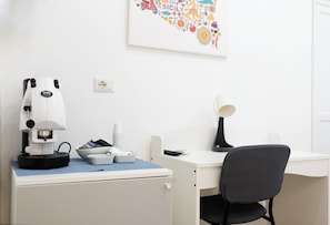Ein schlichter, moderner Raum mit weißem Schreibtisch, schwarzem Bürostuhl, einer Kaffeemaschine und einer Schreibtischlampe.
