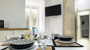 A dining area with a table set for four, a television mounted on the wall, and a view of the outdoors through a window. - ELORO - Guest Houses (Noto)