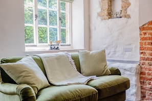 Ein grünes Sofa mit gemustertem Überwurf, ein Fenster mit Blick auf Grünflächen und eine Ziegelwand mit einem Kandelaber.