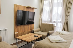 A hotel room with a flat-screen TV mounted on a wooden panel, a beige sofa, a small wooden table with a remote control, and a neatly made bed with folded towels.