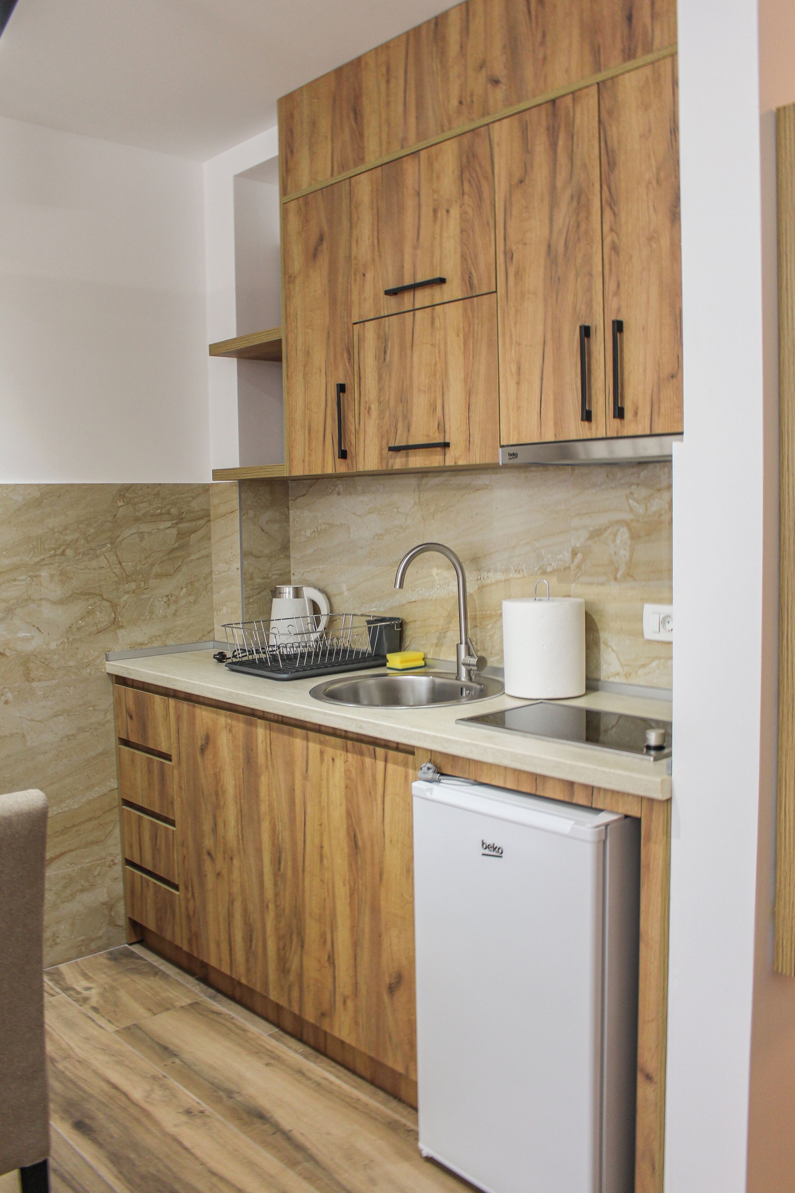 A compact kitchen with wooden cabinets, a built-in sink, a kettle, and a small refrigerator.