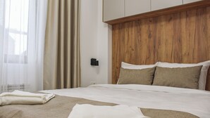 A hotel room with a wooden headboard, a neatly made bed with white linens, and a window with sheer curtains. - Mountain Bliss Spa Resort – Luxury Apartments with Sauna & Jacuzzi (Kolasin)