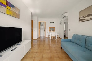 Ein Wohnzimmer mit Fernseher, einem blauen Sofa, einem Essbereich mit Tisch und Stühlen sowie einer Küche im Hintergrund.