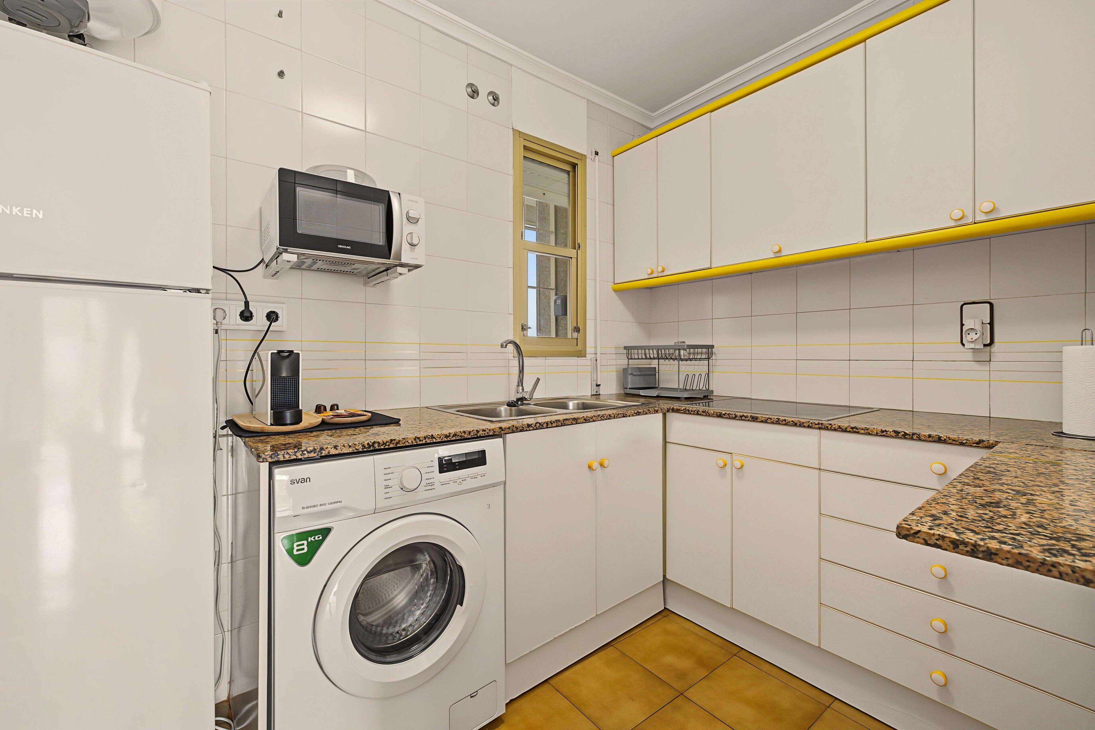 A compact kitchen with white cabinets, a built-in microwave, a washing machine, and a granite countertop.