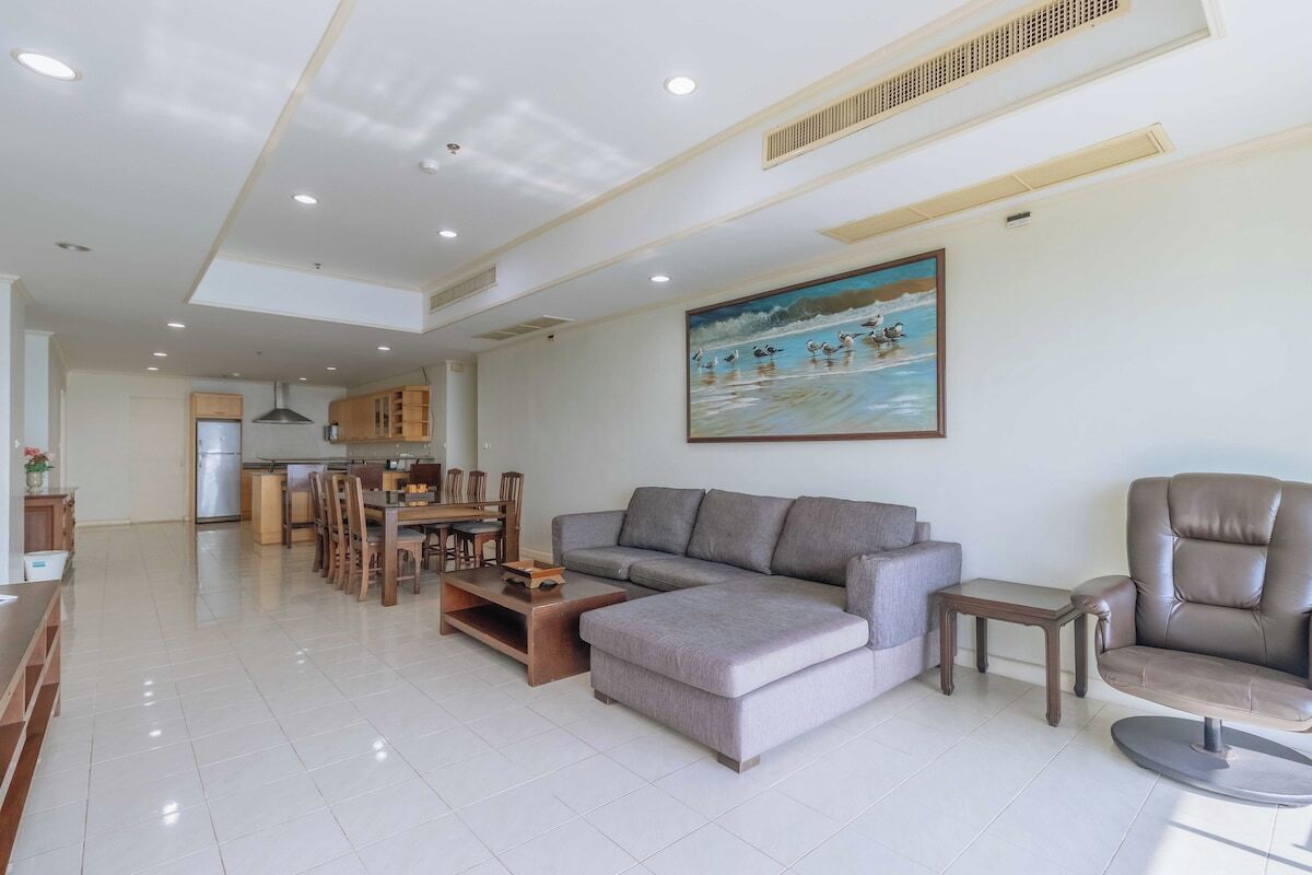 A spacious living area with a grey sectional sofa, a brown leather chair, a dining table with chairs, and a painting of birds on water.